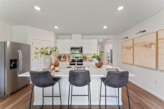 kitchen with light countertops, appliances with stainless steel finishes, white cabinetry, an island with sink, and wood finished floors