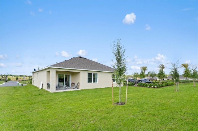 back of house with a shingled roof, stucco siding, a yard, and a patio