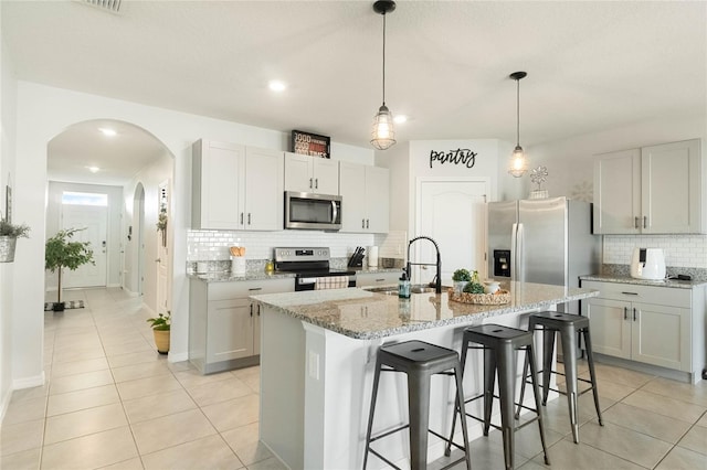 kitchen with arched walkways, a kitchen island with sink, stainless steel appliances, a sink, and light stone countertops