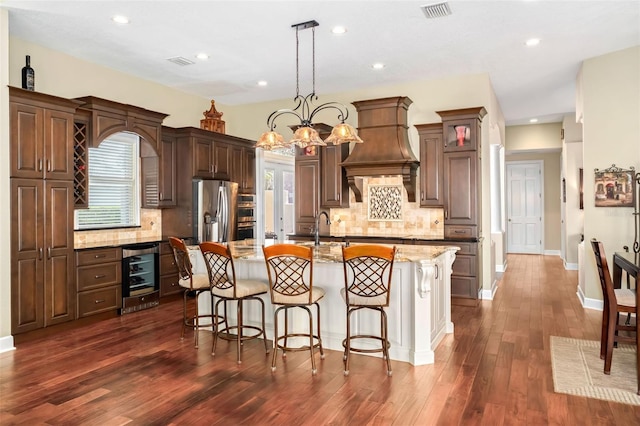 kitchen featuring light stone counters, stainless steel refrigerator with ice dispenser, hanging light fixtures, an island with sink, and beverage cooler