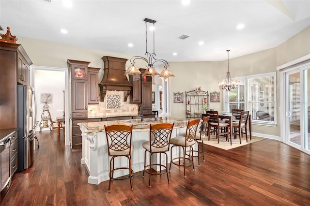 kitchen with a center island with sink, light stone counters, freestanding refrigerator, decorative light fixtures, and premium range hood