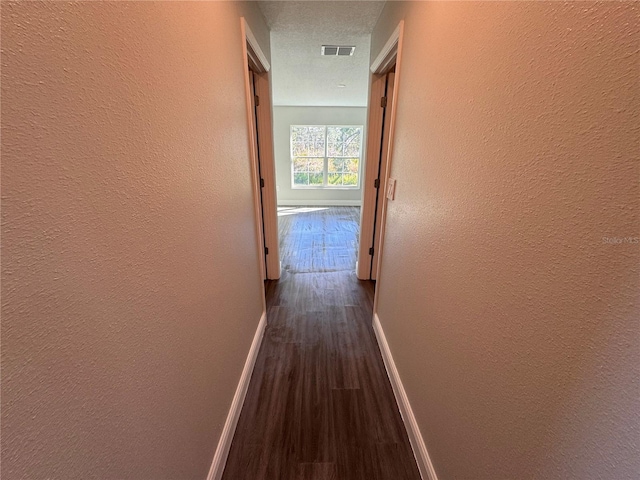 hall featuring dark wood-style floors, a textured wall, visible vents, and baseboards