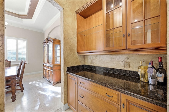 kitchen featuring baseboards, dark stone counters, glass insert cabinets, brown cabinets, and ornamental molding