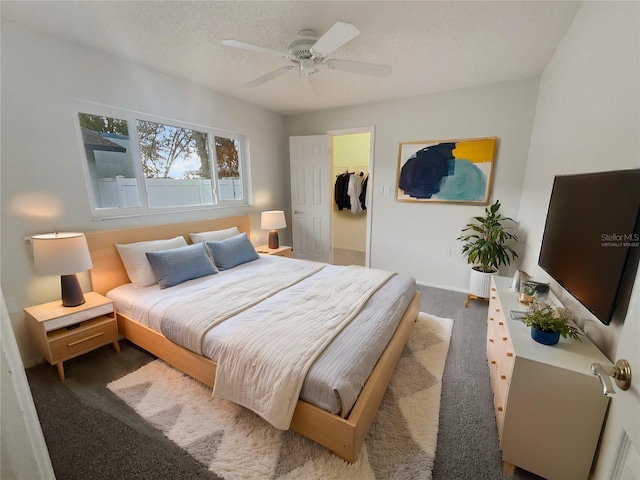 bedroom with a textured ceiling, a ceiling fan, baseboards, dark colored carpet, and a walk in closet