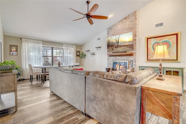 living area with ceiling fan, high vaulted ceiling, visible vents, light wood-style floors, and a brick fireplace