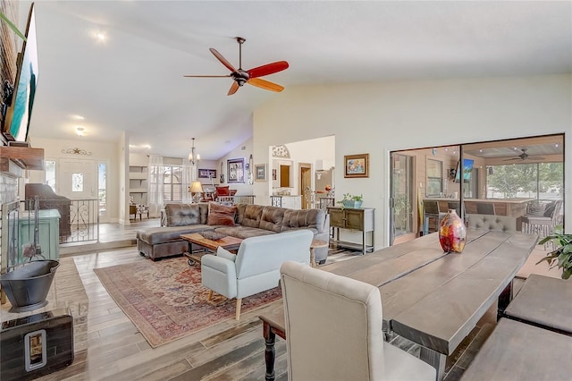 dining room with light wood finished floors, high vaulted ceiling, a fireplace with raised hearth, and ceiling fan with notable chandelier