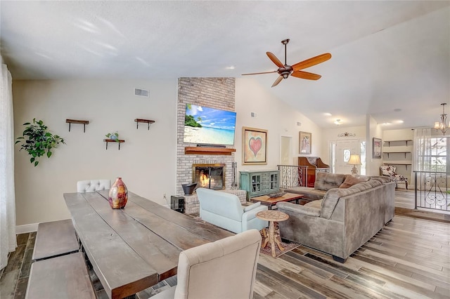 living area with visible vents, light wood-style flooring, a brick fireplace, high vaulted ceiling, and ceiling fan with notable chandelier