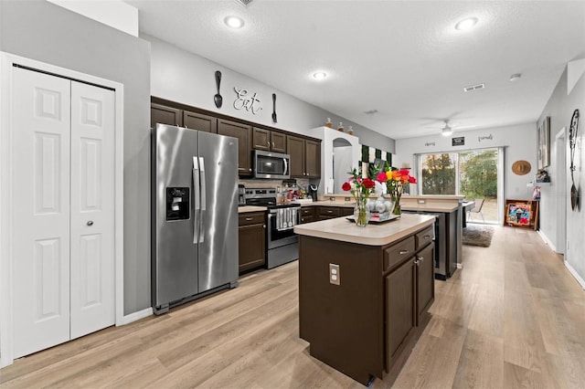 kitchen with light countertops, appliances with stainless steel finishes, a kitchen island, dark brown cabinets, and light wood-type flooring