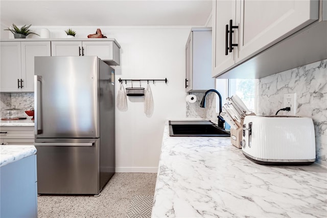 kitchen with light stone counters, decorative backsplash, freestanding refrigerator, a sink, and baseboards