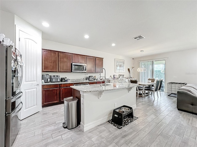 kitchen with visible vents, hanging light fixtures, appliances with stainless steel finishes, a kitchen island with sink, and a sink