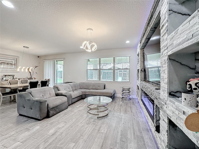 living area featuring light wood finished floors, baseboards, a textured ceiling, a stone fireplace, and a notable chandelier