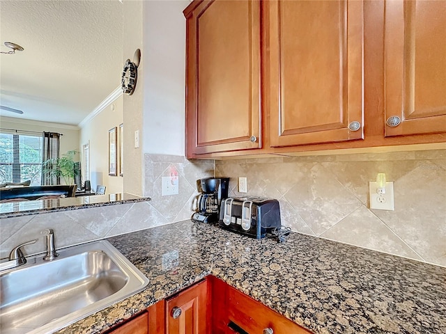 kitchen featuring tasteful backsplash, ornamental molding, brown cabinets, dark stone countertops, and a sink