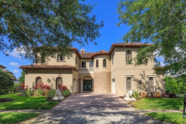 mediterranean / spanish house featuring decorative driveway, stucco siding, a balcony, a tiled roof, and a front lawn