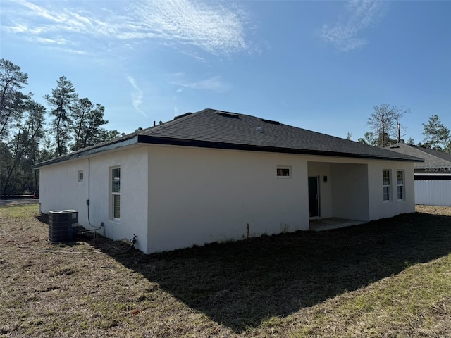 rear view of property featuring stucco siding, a lawn, cooling unit, and roof with shingles