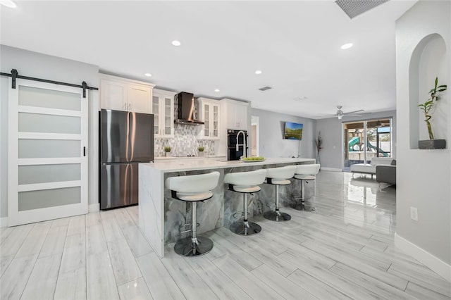 kitchen with a barn door, a kitchen island with sink, light countertops, freestanding refrigerator, and wall chimney exhaust hood