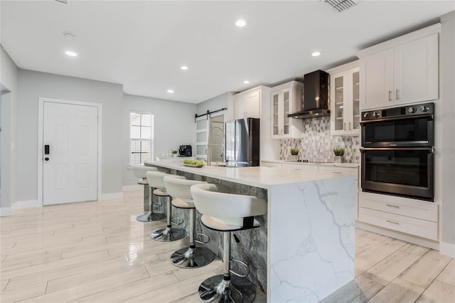 kitchen with double wall oven, a barn door, freestanding refrigerator, white cabinetry, and wall chimney exhaust hood