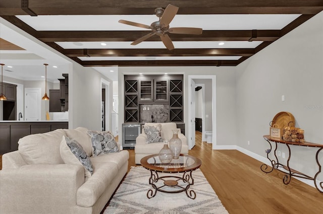 living area featuring baseboards, a ceiling fan, light wood-style flooring, a bar, and beam ceiling