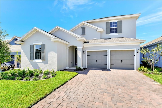 traditional-style home with decorative driveway, stucco siding, a shingled roof, an attached garage, and a front lawn