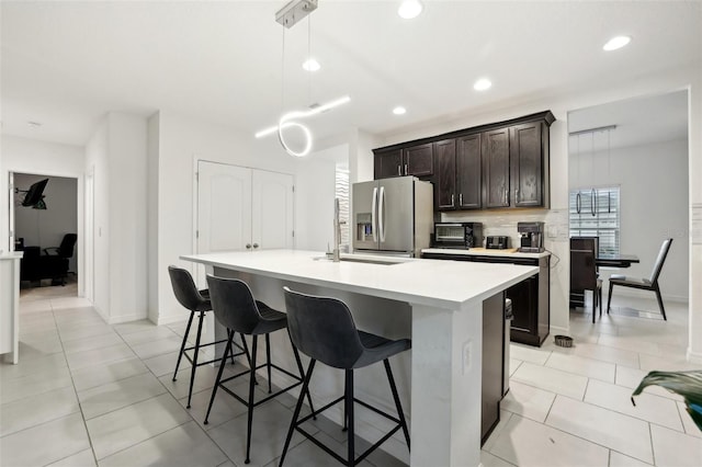 kitchen with stainless steel fridge, an island with sink, decorative light fixtures, light countertops, and dark brown cabinets