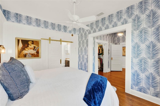 bedroom with a barn door, connected bathroom, wood finished floors, visible vents, and wallpapered walls