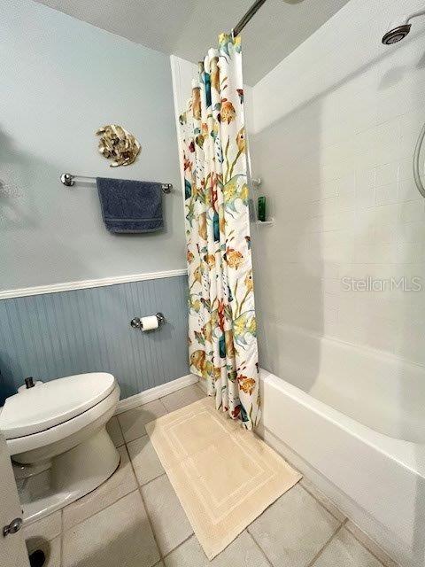 bathroom featuring a wainscoted wall, shower / bathtub combination with curtain, toilet, and tile patterned floors