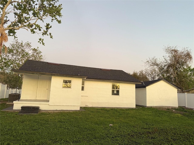 rear view of house featuring a shingled roof, central AC unit, fence, a yard, and stucco siding