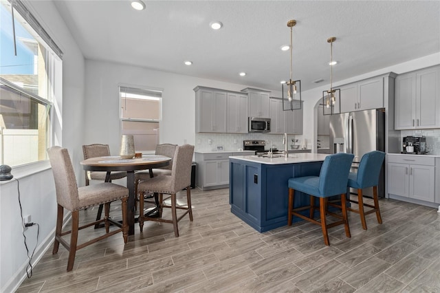 kitchen with gray cabinetry, stainless steel appliances, light countertops, a center island with sink, and pendant lighting