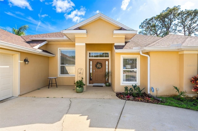 view of exterior entry featuring an attached garage, roof with shingles, and stucco siding