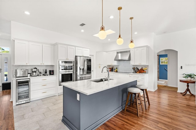 kitchen featuring wine cooler, pendant lighting, stainless steel appliances, white cabinetry, and wall chimney range hood