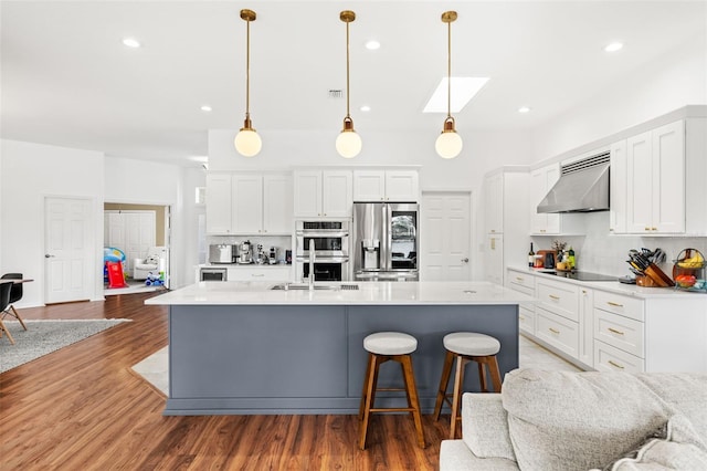 kitchen with hanging light fixtures, appliances with stainless steel finishes, white cabinets, wall chimney range hood, and an island with sink