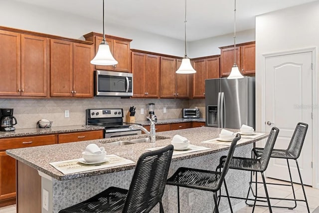 kitchen featuring brown cabinetry, appliances with stainless steel finishes, a breakfast bar, hanging light fixtures, and a kitchen island with sink