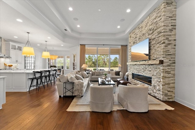 living area featuring a stone fireplace, recessed lighting, baseboards, dark wood-style floors, and a tray ceiling