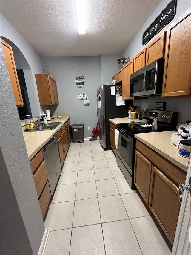 kitchen featuring a sink, a textured ceiling, stainless steel appliances, light tile patterned flooring, and brown cabinetry