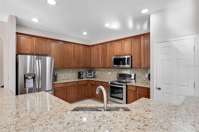 kitchen with decorative backsplash, light stone counters, brown cabinets, stainless steel appliances, and a sink