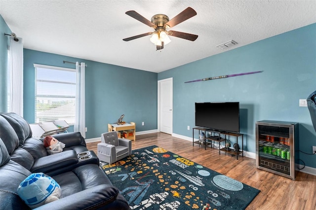 living area featuring ceiling fan, wood finished floors, a textured ceiling, baseboards, and wine cooler