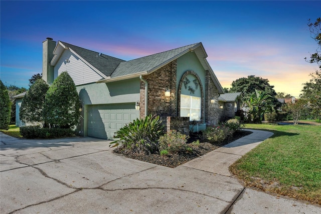 property exterior at dusk featuring brick siding, a chimney, a lawn, an attached garage, and driveway