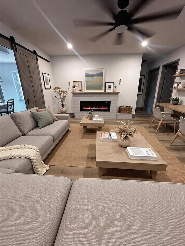 living area with a warm lit fireplace, a barn door, recessed lighting, wood finished floors, and a ceiling fan