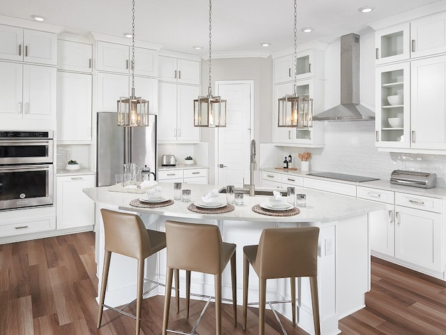 kitchen featuring a kitchen island with sink, wall chimney exhaust hood, appliances with stainless steel finishes, and glass insert cabinets