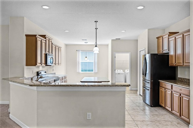 kitchen featuring washing machine and dryer, stainless steel appliances, dark stone counters, brown cabinetry, and decorative light fixtures