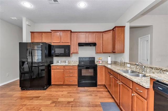 kitchen featuring light wood-style flooring, light stone counters, under cabinet range hood, black appliances, and a sink