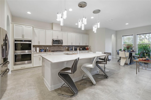 kitchen with a center island with sink, white cabinets, hanging light fixtures, stainless steel appliances, and light countertops