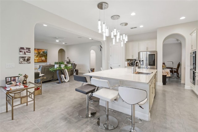 kitchen with arched walkways, a kitchen island with sink, a sink, light countertops, and stainless steel refrigerator with ice dispenser