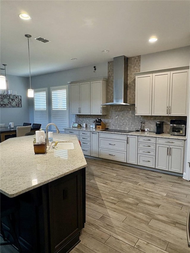 kitchen with black electric cooktop, a sink, wall chimney range hood, light wood finished floors, and tasteful backsplash