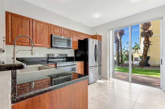 kitchen featuring light tile patterned flooring, stainless steel appliances, a sink, brown cabinets, and dark stone counters