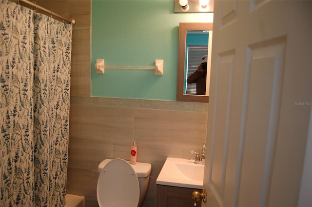 bathroom with toilet, a wainscoted wall, a tile shower, vanity, and tile walls