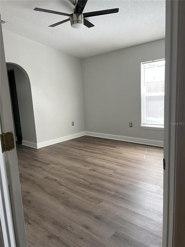 empty room featuring a ceiling fan, a textured ceiling, dark wood finished floors, arched walkways, and baseboards