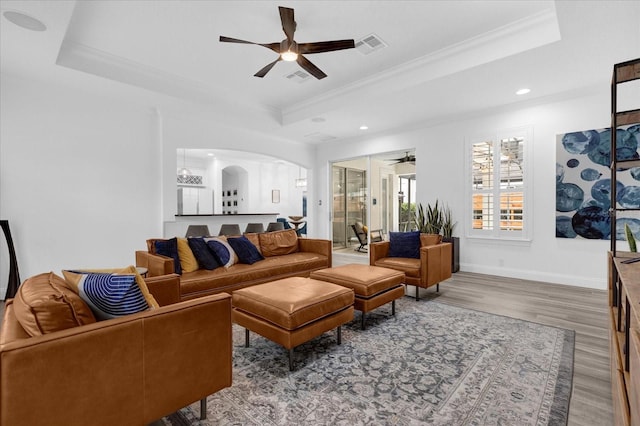 living area with ceiling fan, arched walkways, wood finished floors, ornamental molding, and a raised ceiling