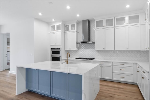 kitchen featuring double oven, light wood-style floors, white cabinets, wall chimney exhaust hood, and a center island with sink