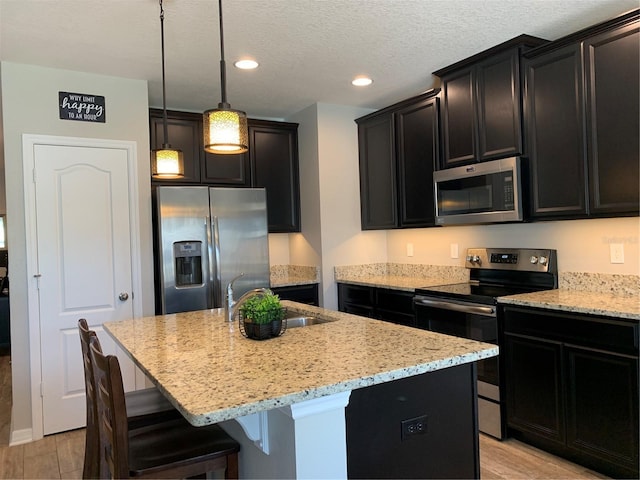 kitchen featuring a center island with sink, stainless steel appliances, light stone countertops, and dark cabinetry