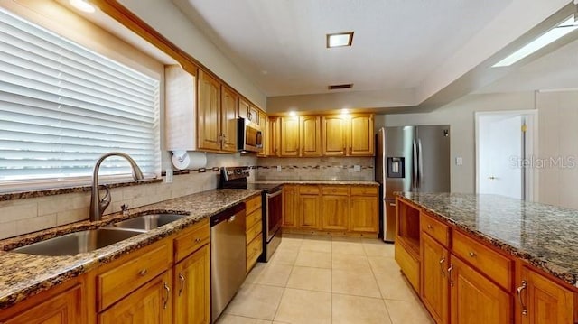 kitchen featuring decorative backsplash, dark stone counters, brown cabinets, stainless steel appliances, and a sink